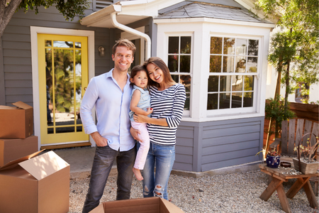 An adult man, adult woman, and young girl stand outside a home with moving boxes, smiling. The woman holds the little girl.